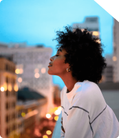 Mulher apreciando a vista da cidade ao entardecer Mulher com cabelo cacheado, usando uma blusa branca, apreciando o horizonte urbano ao entardecer, com prédios ao fundo e luzes da cidade iluminando o cenário.