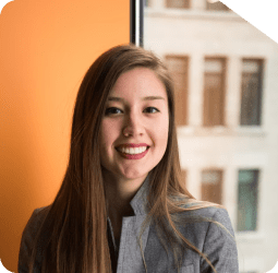 Mulher sorridente com fundo urbano Mulher com cabelos castanhos longos, usando blazer cinza, sorrindo para a câmera com uma janela ao fundo que mostra a fachada de um edifício em tons claros.