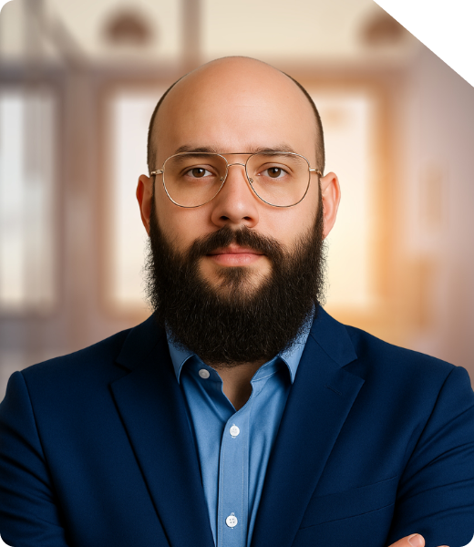 Marcius, CEO da Shinier Marcius, CEO da Shinier, em retrato profissional. Ele está com a cabeça raspada, usa barba cheia bem desenhada e óculos de armação dourada estilo aviador. Veste uma camisa social azul claro sob um blazer azul escuro, transmitindo profissionalismo e confiança. O fundo está desfocado, com luz natural suave entrando por janelas, sugerindo um ambiente corporativo moderno.