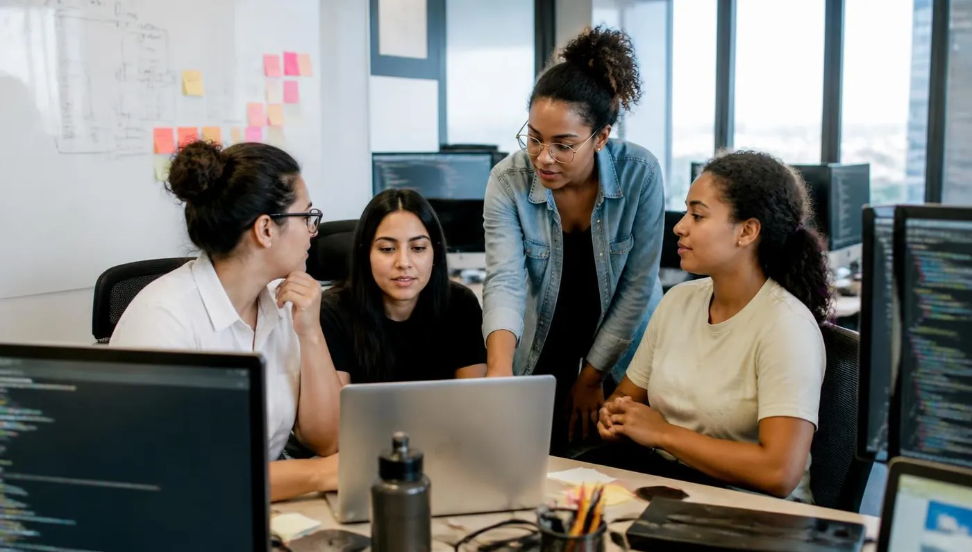 Quatro mulheres profissionais de tecnologia trabalhando juntas em frente a um notebook em um escritório de startup