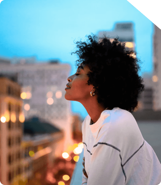 Mulher com cabelo cacheado, usando uma blusa branca, apreciando o horizonte urbano ao entardecer, com prédios ao fundo e luzes da cidade iluminando o cenário.