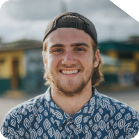 Homem jovem usando camisa estampada e boné para trás, sorrindo para a câmera em um ambiente ao ar livre com edifícios desfocados ao fundo.