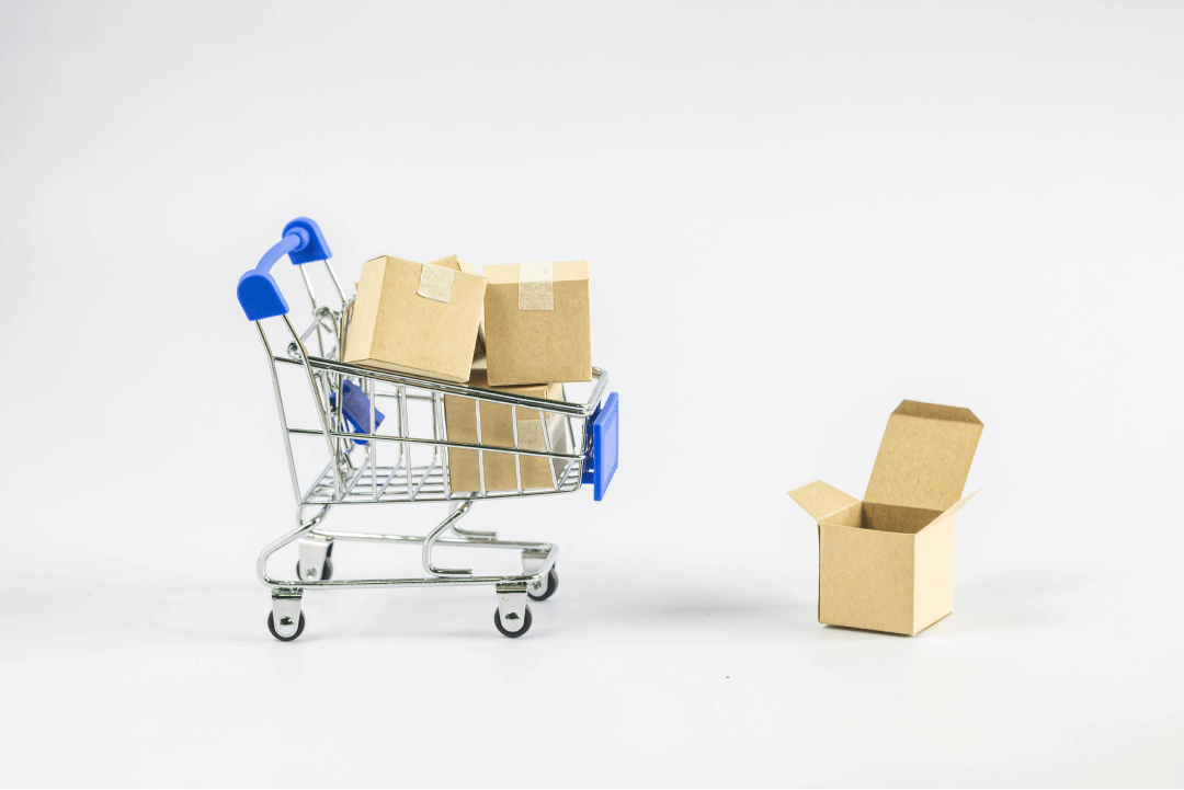 A imagem mostra um pequeno carrinho de compras de metal com detalhes em azul, contendo várias caixas de papelão, simbolizando compras ou logística de e-commerce. Ao lado do carrinho, há uma caixa aberta, sugerindo a preparação para embalagem ou envio de mercadorias. O fundo branco minimalista destaca os itens no carrinho, reforçando a ideia de um marketplace online, compras digitais ou processos de envio e entrega de produtos. O cenário sugere simplicidade e clareza, representando o conceito de comércio eletrônico e gestão de pedidos.