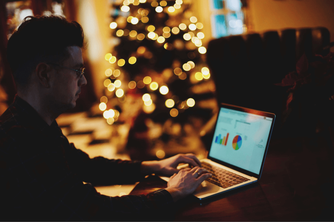 A imagem mostra uma pessoa usando um laptop, aparentemente concentrada em trabalhar ou analisar gráficos que estão visíveis na tela. No fundo, há uma árvore de Natal decorada com luzes brilhantes, criando uma atmosfera aconchegante e festiva. As luzes desfocadas ao fundo contrastam com o foco no trabalho, sugerindo um equilíbrio entre a época festiva e o comprometimento com as atividades profissionais. O ambiente parece ser um espaço doméstico ou de home office, destacando o cenário de trabalho remoto.