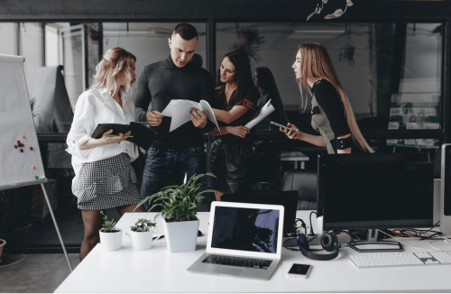 A imagem mostra um grupo de quatro pessoas em um ambiente de escritório moderno, colaborando e discutindo documentos. Eles estão de pé em frente a uma mesa, que possui laptops, fones de ouvido, plantas e outros itens de trabalho. O quadro branco ao fundo sugere um ambiente voltado para brainstorming ou planejamento. O grupo parece estar engajado em uma conversa produtiva, com duas mulheres e um homem no centro da imagem, revisando papéis juntos, enquanto outra mulher à direita segura um smartphone. O cenário sugere uma equipe jovem e dinâmica trabalhando em conjunto.