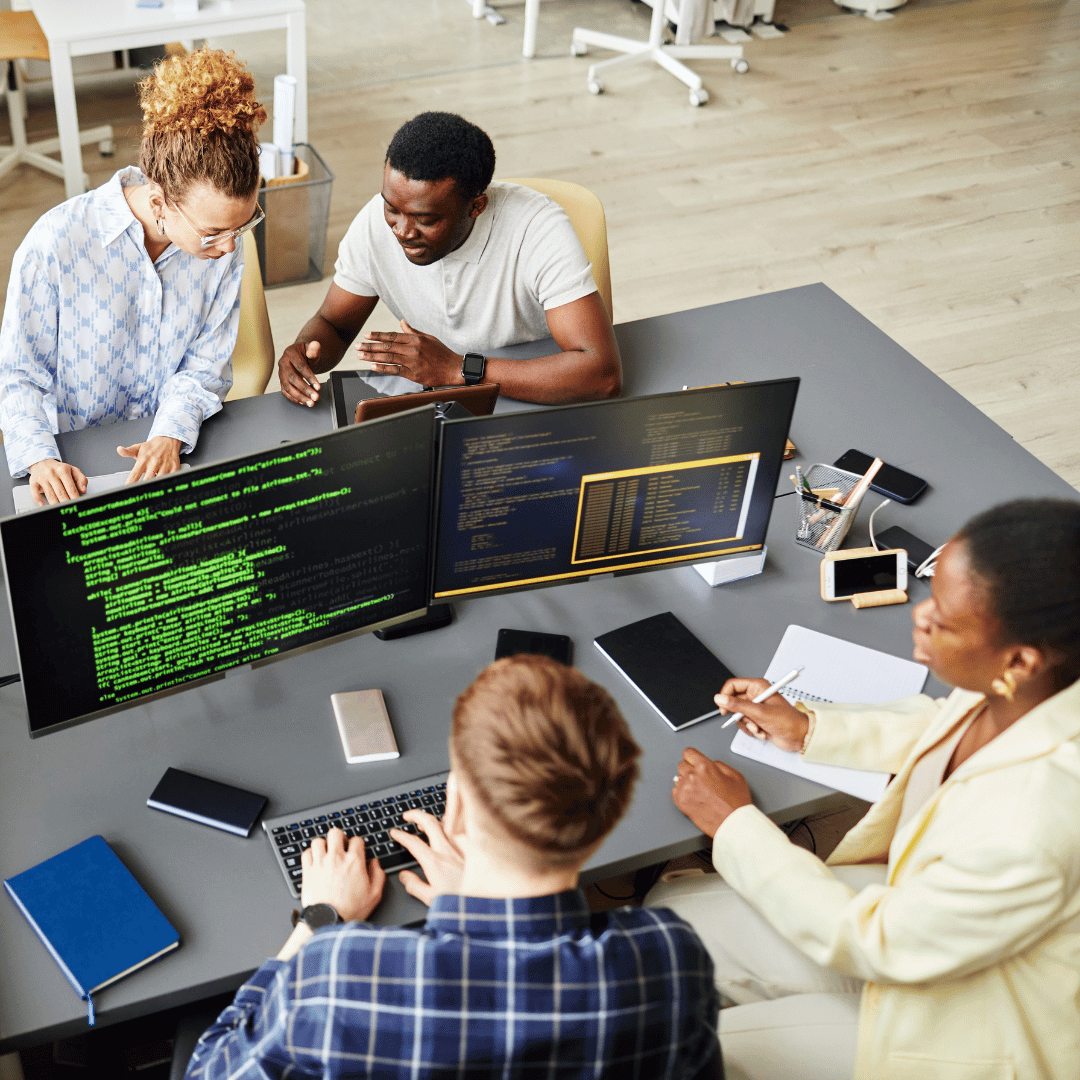 Grupo de quatro pessoas trabalhando em equipe em torno de uma mesa com dois monitores mostrando código de programação. Três pessoas estão interagindo e discutindo, enquanto uma delas está no computador digitando. Diversos dispositivos, como celulares e notebooks, estão espalhados pela mesa. O ambiente parece ser um escritório moderno.