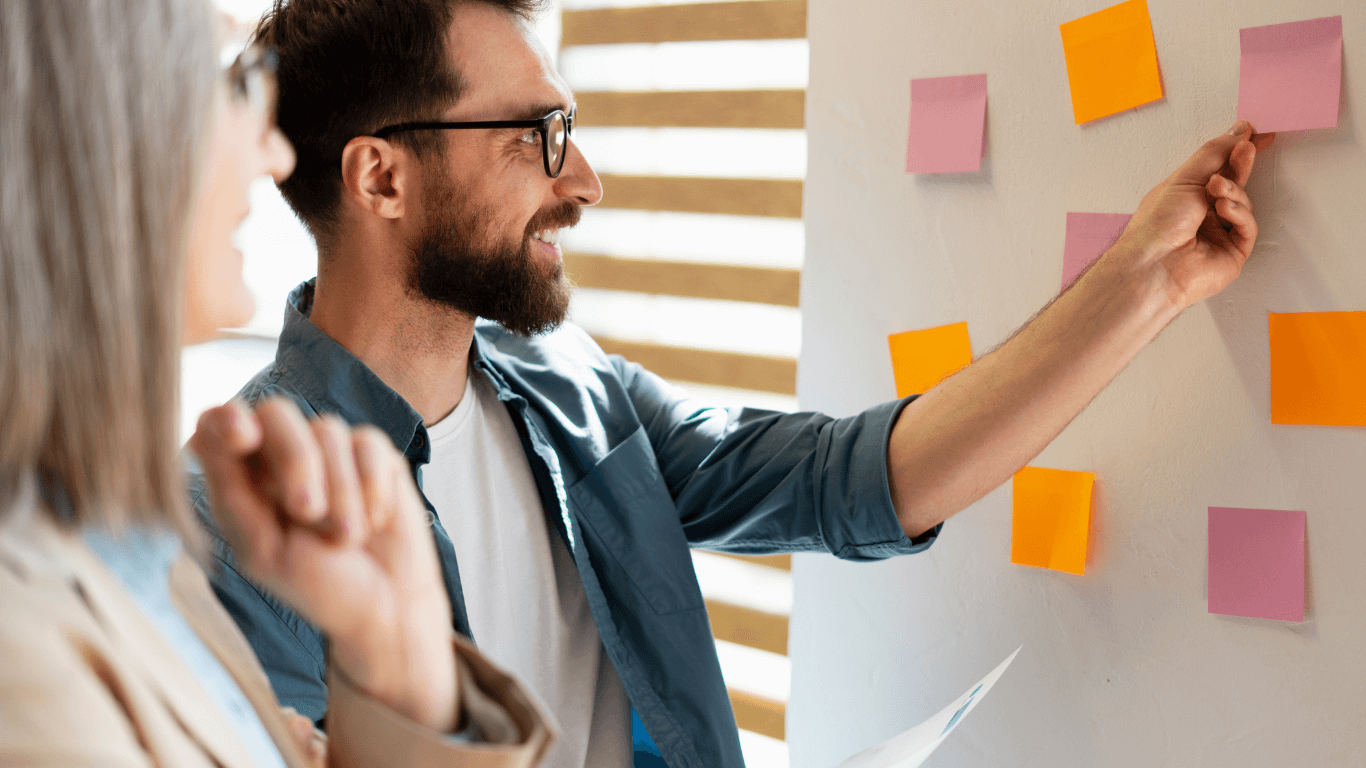 Homem sorridente organizando post-its coloridos em uma parede, colaborando em uma atividade de planejamento de projeto em equipe, enquanto outra pessoa observa atentamente.