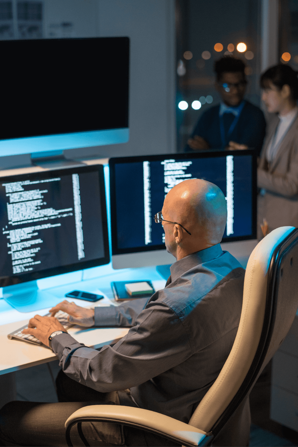 Homem maduro e calvo usando óculos, focado na análise de dados e programação em uma estação de trabalho com vários monitores. Na tela, ele observa linhas de código e visualizações digitais em um ambiente moderno e iluminado, enquanto duas outras pessoas discutem ao fundo.