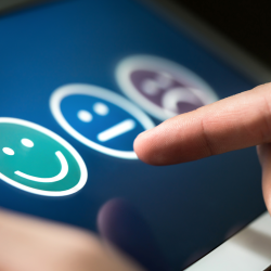 Feedback Dedos tocando a tela de um tablet para selecionar um ícone de satisfação representado por um rosto sorridente. A interface apresenta opções de feedback visual, incluindo expressões neutras e insatisfeitas, indicando um sistema de avaliação digital.