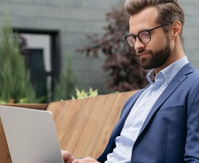 Homem de terno azul, sentado em um banco de madeira, trabalhando em um laptop em um ambiente externo com vegetação ao fundo.