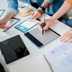 Ajustes Equipe de designers e desenvolvedores trabalhando na criação de um MVP, utilizando uma mesa digitalizadora e analisando wireframes e protótipos de interface de usuário. No ambiente de trabalho, há tablets, anotações e esboços de design, destacando o processo criativo de desenvolvimento de produtos digitais.