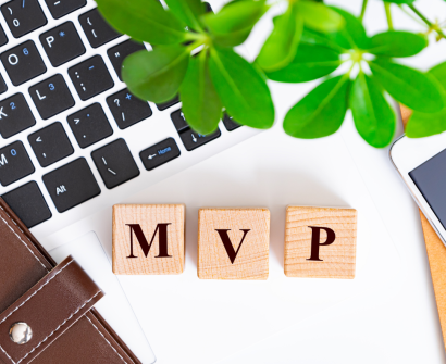 Blocos de madeira com as letras "MVP" sobre um teclado de notebook, simbolizando o conceito de Minimum Viable Product (Produto Mínimo Viável). Ao redor, elementos como uma carteira de couro, um smartphone e folhas verdes remetem à inovação e desenvolvimento de negócios.