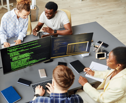 Grupo de quatro pessoas trabalhando em equipe em torno de uma mesa com dois monitores mostrando código de programação. Três pessoas estão interagindo e discutindo, enquanto uma delas está no computador digitando. Diversos dispositivos, como celulares e notebooks, estão espalhados pela mesa. O ambiente parece ser um escritório moderno.